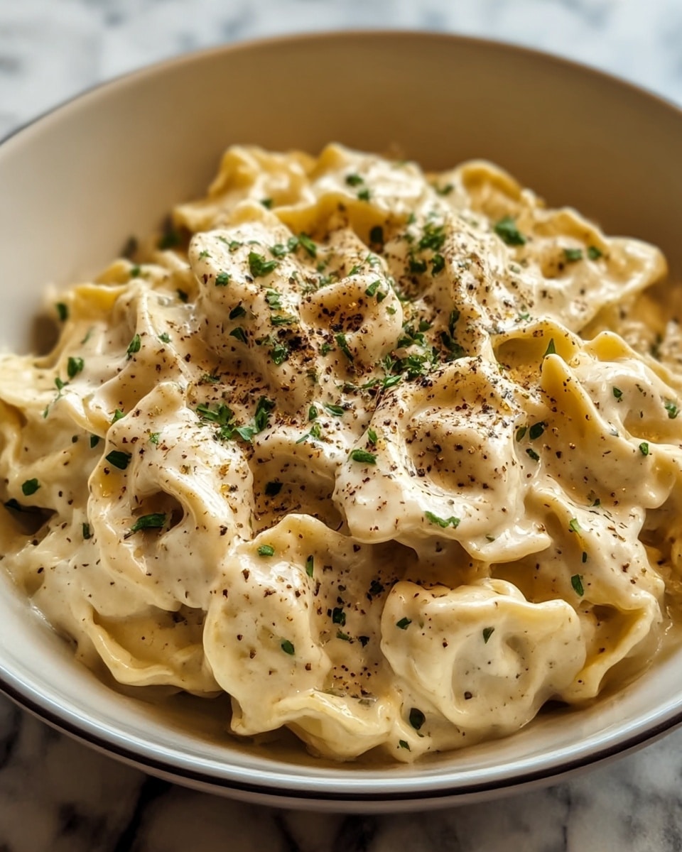A bowl filled with a creamy pasta dish consisting of tortellini coated in a thick, smooth, off-white sauce. The tortellini pieces are arranged closely together, showing slightly folded shapes with soft, delicate edges. The dish is topped with chopped green herbs scattered lightly on top and freshly ground black pepper, adding specks of dark brown contrast across the creamy surface. The bowl is white with a simple design, and the background is a white marbled texture. photo taken with an iphone --ar 4:5 --v 7