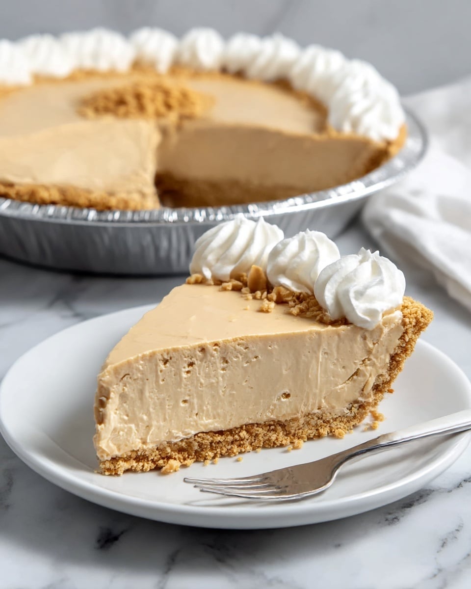 A slice of peanut butter pie sits on a white round plate, showing three layers: a crumbly golden brown crust at the bottom, a thick light tan creamy filling in the middle, and a smooth pale tan top layer. The edge of the slice is decorated with three white swirls of whipped cream. In the background, the rest of the pie is in a shiny silver pie tin, with a similar whipped cream border around the edge and a small cluster of crushed nuts in the center. A silver fork with a white handle rests near the pie slice on the plate, all placed on a white marbled surface. Photo taken with an iphone --ar 4:5 --v 7