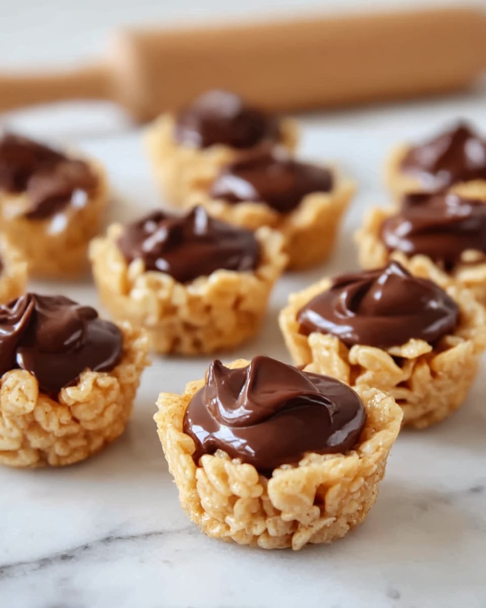 The image shows nine small rice cereal treats shaped like cups arranged on a white marbled surface. Each cup has two layers: the bottom and sides are made of golden, crispy rice cereal held together, forming a rough textured cup shape. The top layer is a smooth, shiny dollop of dark brown chocolate filling, slightly glossy and thick, sitting inside each rice cereal cup. The cups are placed close together, and the background is softly blurred with a rolling pin visible behind. Photo taken with an iphone --ar 4:5 --v 7