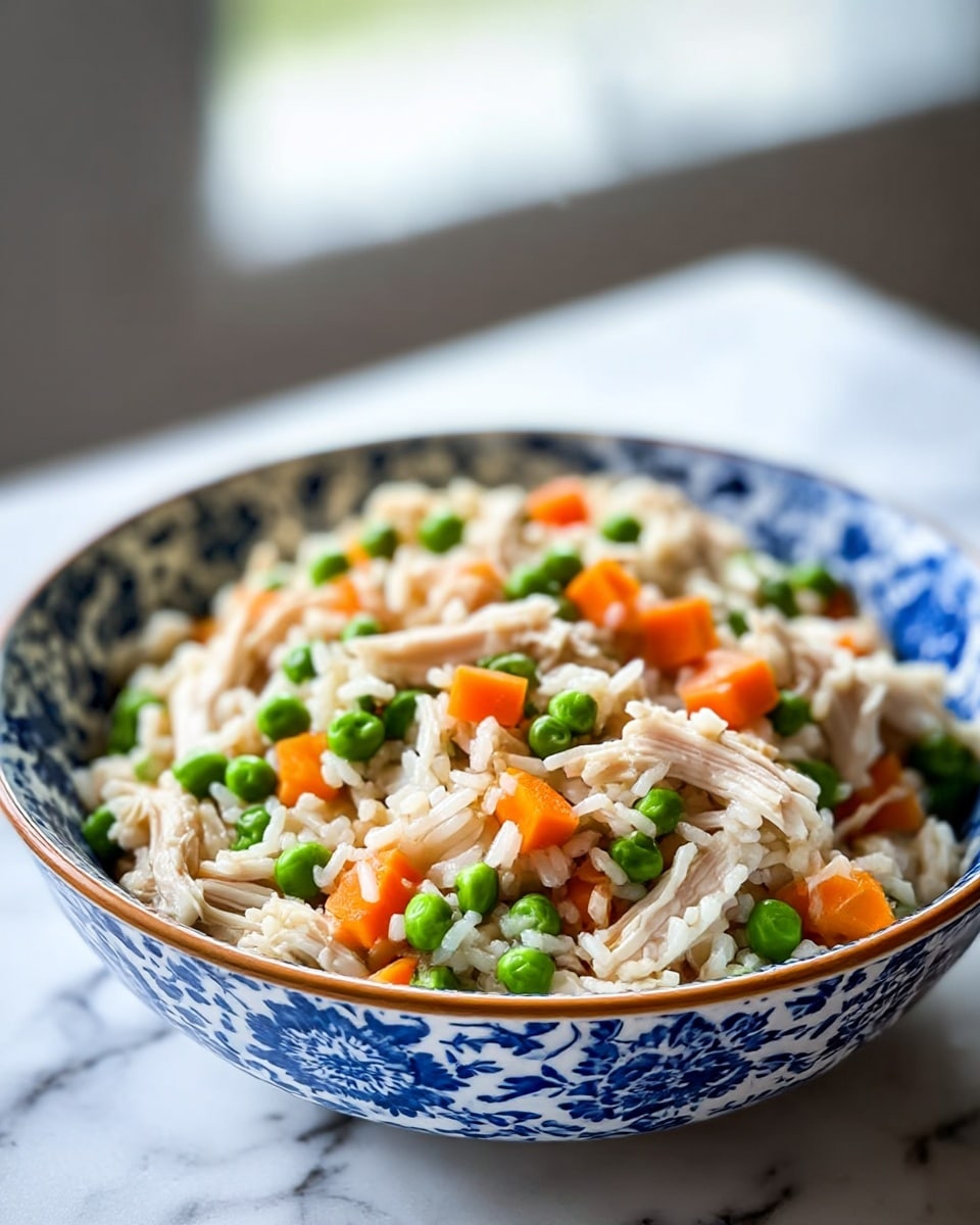 A white bowl with blue leafy patterns holds a colorful rice dish with three main layers visible: the base layer is light brown, fluffy cooked rice mixed evenly with bright green peas and soft orange carrot cubes, and the top layer consists of shredded, pale cooked chicken pieces scattered over the rice and vegetables. The bowl is placed on a white marbled surface with a blurred background featuring a green potted plant and a yellow bottle. The scene is softly lit with natural light. photo taken with an iphone --ar 4:5 --v 7