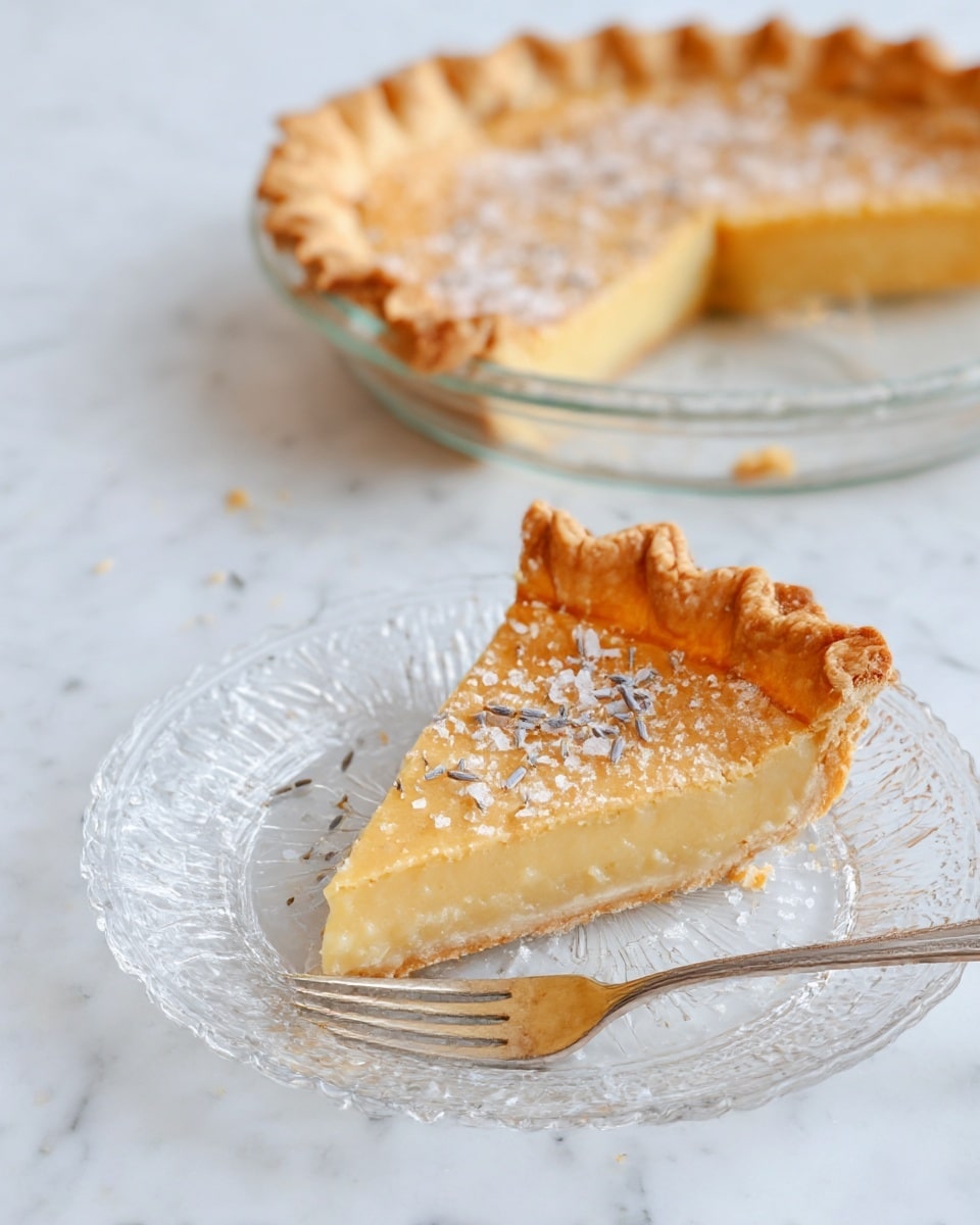 The image shows a slice of pie on a clear glass plate with a detailed edge, placed on a white marbled surface. The pie has two visible layers: a golden-brown crust with a crimped edge on top and a pale yellow, smooth filling beneath. The crust is slightly textured and sprinkled with lavender flowers and coarse salt. A small triangular piece of the pie is cut off and sits beside the slice with a silver fork resting on the plate, pointing toward the pie. In the background, the remaining pie is in a clear glass dish, showing the same golden crust and pale yellow filling with coarse salt and lavender on top. photo taken with an iphone --ar 4:5 --v 7