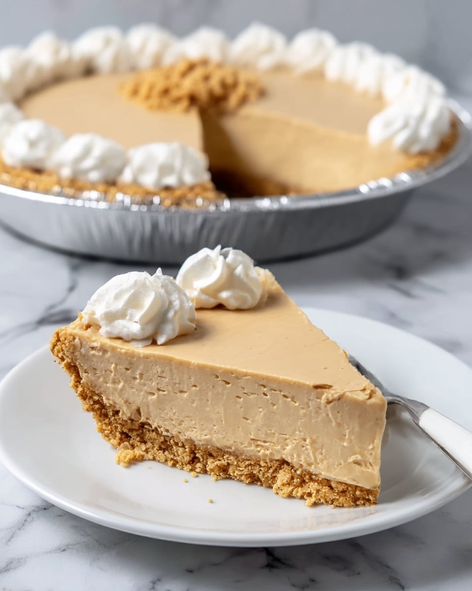 A slice of creamy light brown pie with a thick, crumbly light tan crust sits on a white plate. The pie slice has a smooth, dense texture and is topped with three swirls of white whipped cream along the back edge. Behind it, the remaining pie in a silver foil pie pan shows the same layers and decoration with more whipped cream swirls around the edge and a few crumbs in the middle. A silver fork rests on the plate near the slice. Everything is set on a white marbled surface. photo taken with an iphone --ar 4:5 --v 7