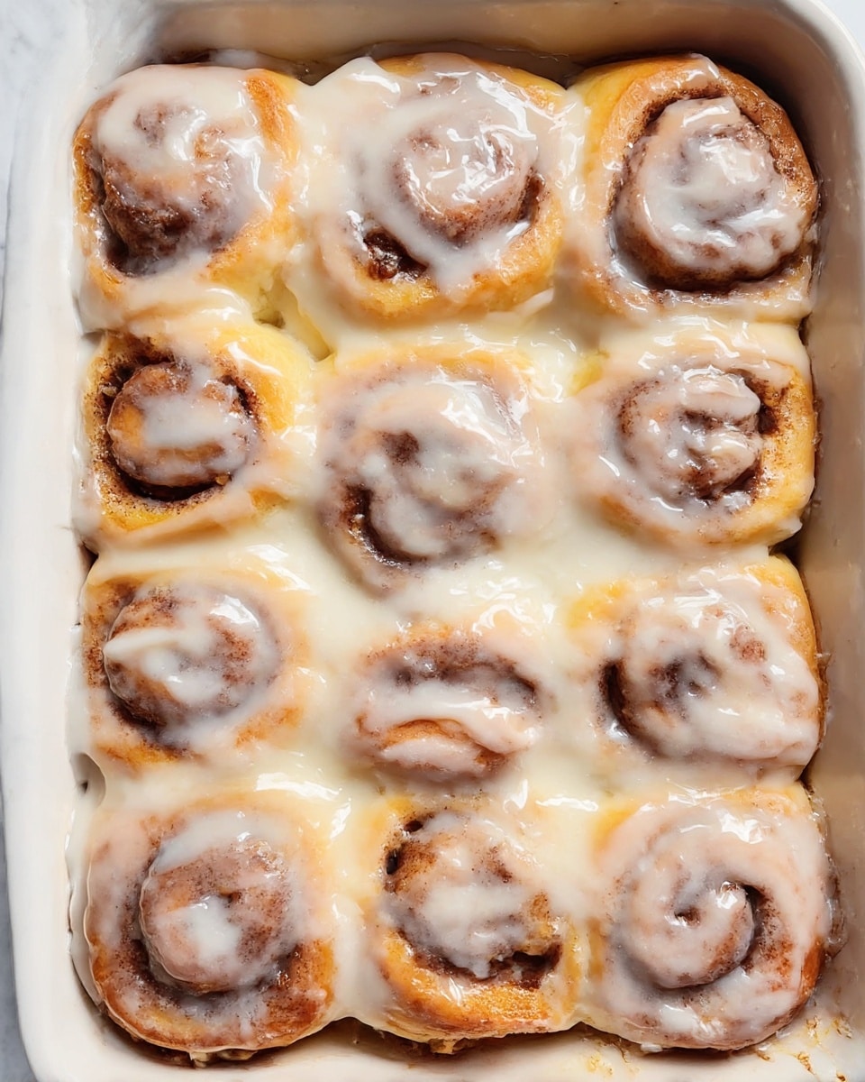The image shows a white baking dish filled with 12 cinnamon rolls arranged closely in three rows of four, each roll featuring a visible spiral of golden-brown dough and dark cinnamon filling. Thick, white icing is generously spread over the top and sides of the rolls, melting slightly and giving a shiny, creamy texture. The rolls are soft and slightly puffed, with some light toasted spots on the top edges. The light reflects subtly on the glossy icing and warm dough. The dish sits on a white marbled surface. Photo taken with an iphone --ar 4:5 --v 7