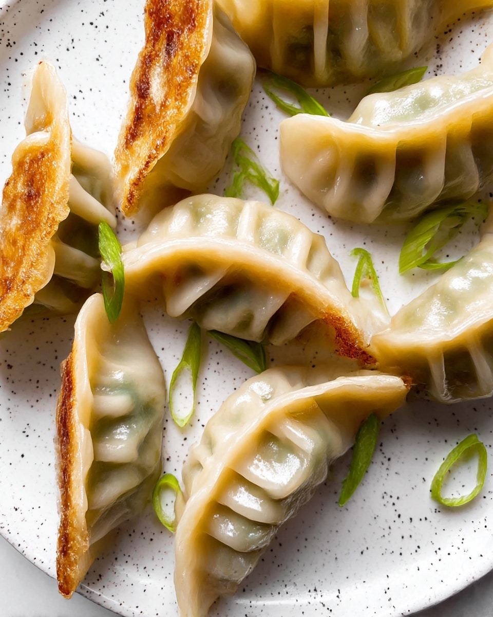 The image shows several pan-fried dumplings arranged closely on a white plate with small black speckles. Each dumpling has a pale, slightly translucent dough wrapper with delicate pleats forming ridges on top, some with a golden brown, crispy side that indicates pan-frying. Visible inside the edges, the filling casts a slight greenish hue through the wrapper. Thin slices of green onion are scattered around the dumplings, adding freshness and a pop of green color. The plate rests on a white marbled surface. photo taken with an iphone --ar 4:5 --v 7