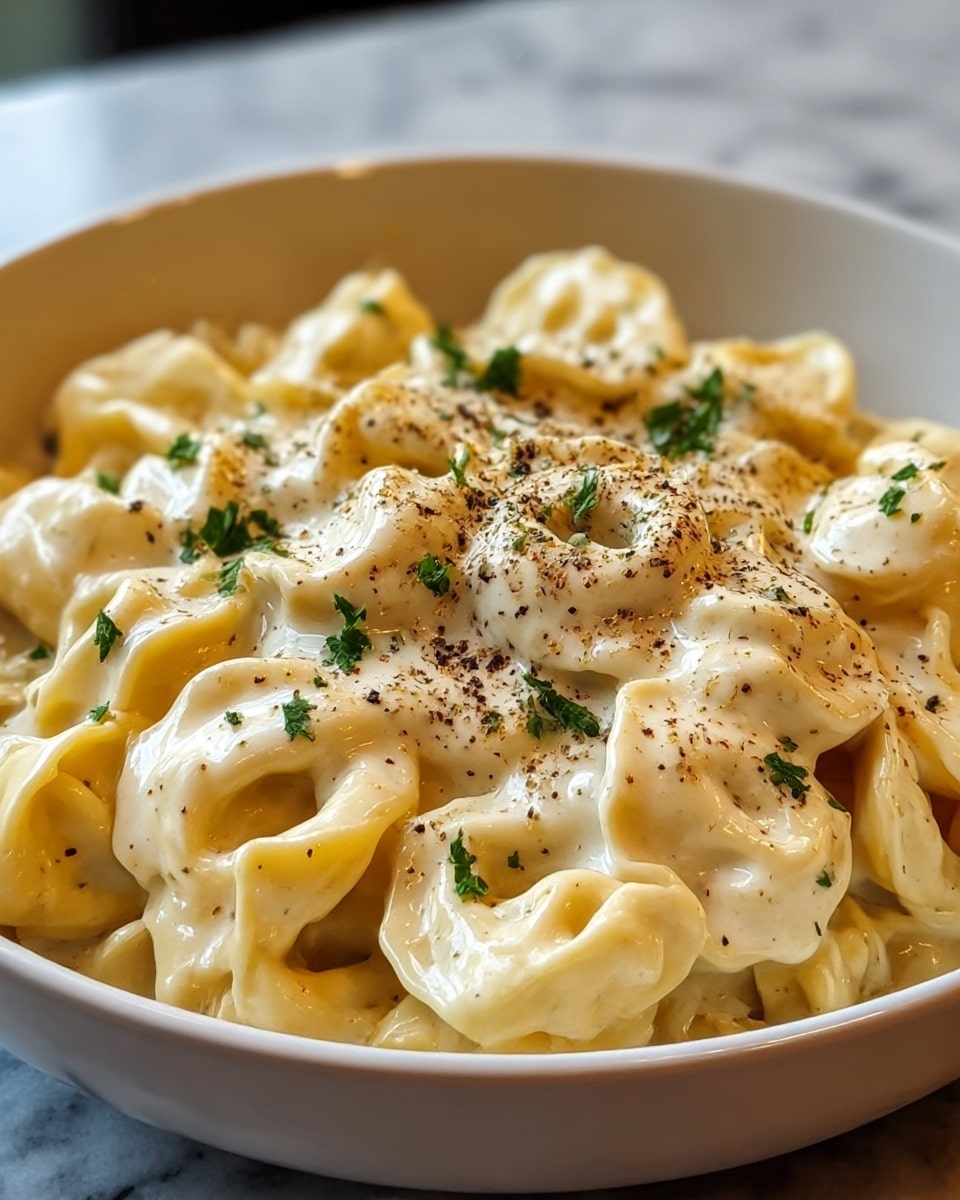 A close-up of a bowl filled with three layers of cheese-filled tortellini pasta, all coated in a creamy, white cheese sauce. The pasta is pale yellow with a smooth texture, and each piece is distinct and rounded. The dish is topped with small green parsley leaves and a sprinkle of black pepper, giving contrast and a touch of freshness. The bowl is white and placed on a white marbled surface. Photo taken with an iphone --ar 4:5 --v 7
