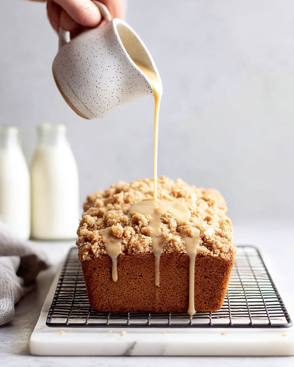 A rectangular loaf of crumb cake with a rich, golden-brown crumb topping that looks crunchy and textured, sitting on a wire rack over a white marble slab; a woman's hand pours a thin stream of creamy, light tan glaze from a small white speckled jug onto the top center of the cake, some glaze dripping down the rough crumb surface; the background shows soft-focus white milk bottles with a white marbled texture under the setup photo taken with an iphone --ar 4:5 --v 7