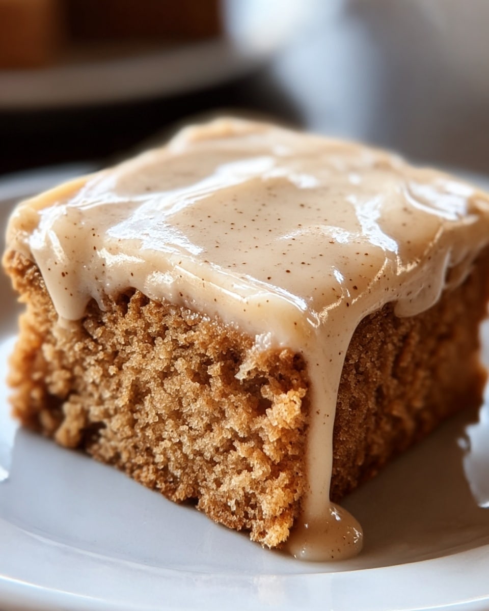 The image shows a close-up of a single square piece of moist cake with a rough texture, light brown in color. It has one thick layer of creamy frosting on top that is tan with tiny darker specks. The frosting looks smooth and slightly glossy, slightly dripping down the sides of the cake. The cake rests on a shiny white plate placed on a white marbled surface. Photo taken with an iphone --ar 4:5 --v 7