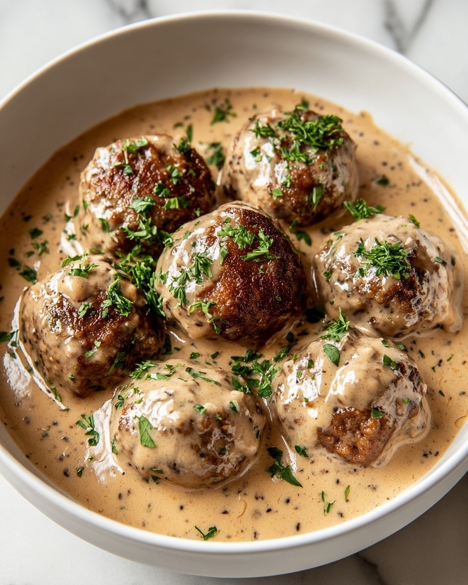 A white bowl filled with seven browned meatballs covered in a thick, creamy light brown sauce with visible pepper specks, garnished with chopped fresh green herbs scattered on top, all placed on a white marbled surface photo taken with an iphone --ar 4:5 --v 7