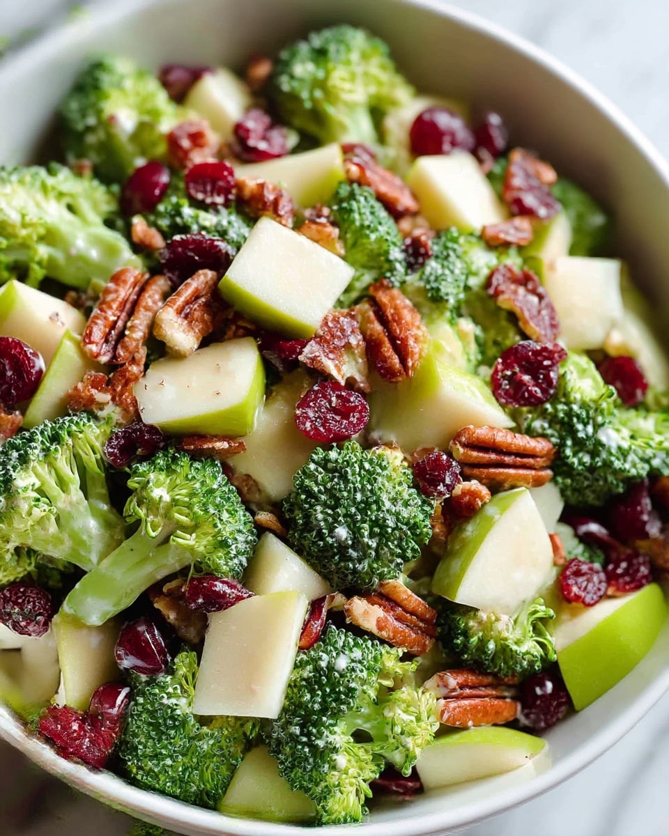A close-up view of a fresh salad in a white bowl placed on a white marbled surface, showing layers of green broccoli florets with a bumpy texture as the base, mixed with chunks of pale green apple pieces, bright red dried cranberries scattered throughout, and brown pecan nuts adding a rough texture all over. The salad appears lightly coated with a creamy dressing lightly visible on some broccoli and nuts. Photo taken with an iphone --ar 4:5 --v 7