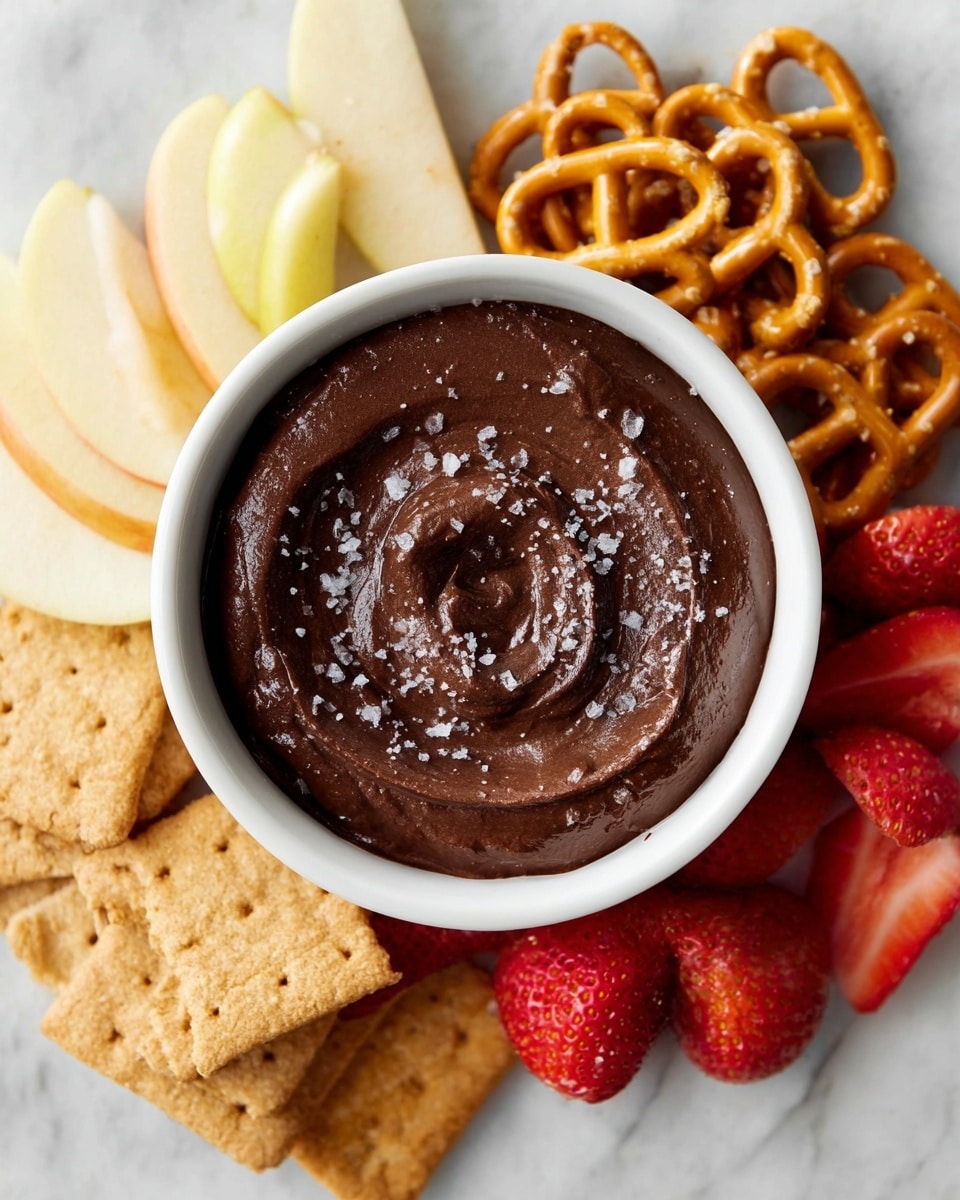 A white bowl filled with thick, dark chocolate dip topped with coarse salt crystals sits in the center, with a silver spoon partially dipped inside, resting on a wooden board. Behind the bowl, on the left side, are slices of pear, pale yellow and layered in a semi-circle. On the right side of the bowl are small, round pretzels, golden brown and crunchy looking, with halved strawberries showing their red juicy interior and green stems beside them. The whole setup is on a white marbled textured surface. photo taken with an iphone --ar 4:5 --v 7
