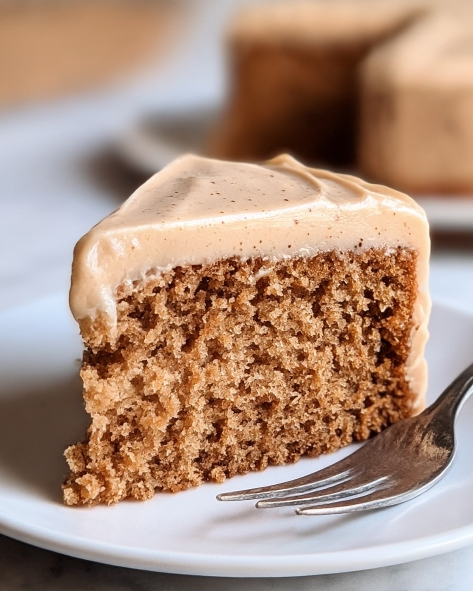 A close-up of a single slice of soft, brown cake with a moist and crumbly texture, topped with a thick layer of smooth, light tan frosting that has tiny darker specks throughout. The cake slice sits on a white plate with part of a metal fork visible nearby. The background shows another blurred slice of cake on a white plate, all set on a white marbled surface. Photo taken with an iphone --ar 4:5 --v 7