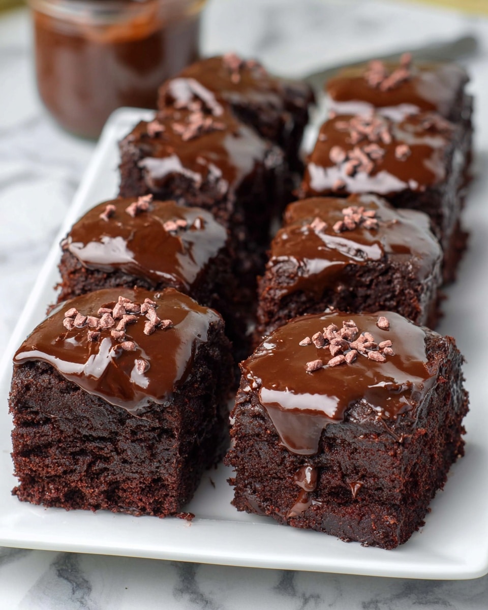 A white rectangular plate holds eight square pieces of rich, dark chocolate cake arranged in two rows. Each piece has thick, glossy dark chocolate frosting spread unevenly on top, with the frosting slightly dripping down the sides. Small chocolate nibs are sprinkled on the glossy frosting pieces, adding texture and contrast. The cake layers are dense and moist, showing a deep brown color with a slightly crumbly texture. The plate sits on a surface with a white marbled texture, and a small glass jar with more chocolate frosting blurred in the background. Photo taken with an iphone --ar 4:5 --v 7