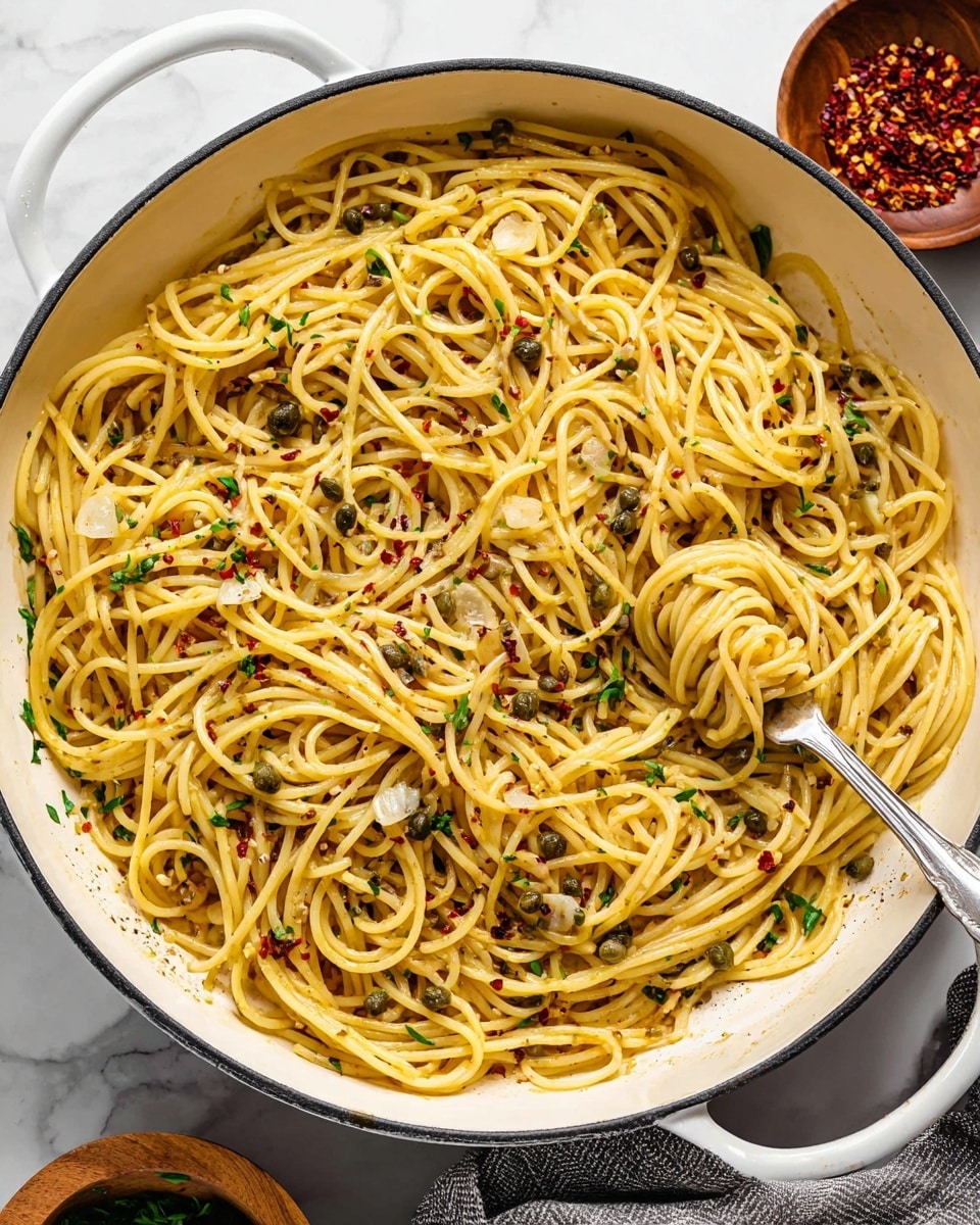A large white pan filled with golden yellow spaghetti noodles twisted in loops, mixed with thin, light beige slices of garlic, small greenish-gray capers, scattered dark green parsley bits, and red chili flakes providing a spicy touch. A silver fork is lifting a small bundle of noodles on the right side of the pan, resting against the dish. The pan is placed on a white marbled surface, with a wooden bowl of red chili flakes and a white bowl with green herbs partially visible. Photo taken with an iphone --ar 4:5 --v 7