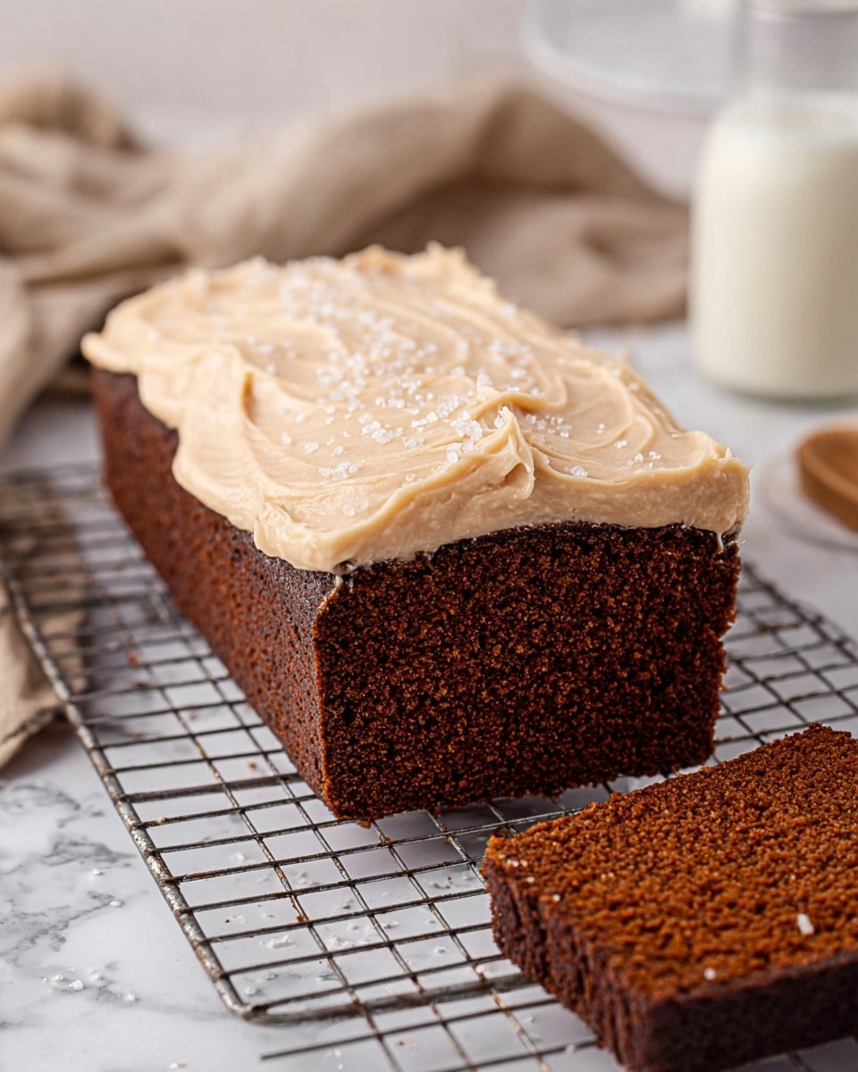 A rectangular dark brown cake with a moist texture sits on a metal cooling rack over a white marbled surface. The cake has one thick layer, topped with a thick smooth creamy light beige frosting that has a swirled texture and is sprinkled lightly with coarse white salt flakes. On the right, a thick slice is cut off, showing the dense, spongy brown inside of the cake. In the blurred background, there is a glass of milk and a beige cloth. photo taken with an iphone --ar 4:5 --v 7