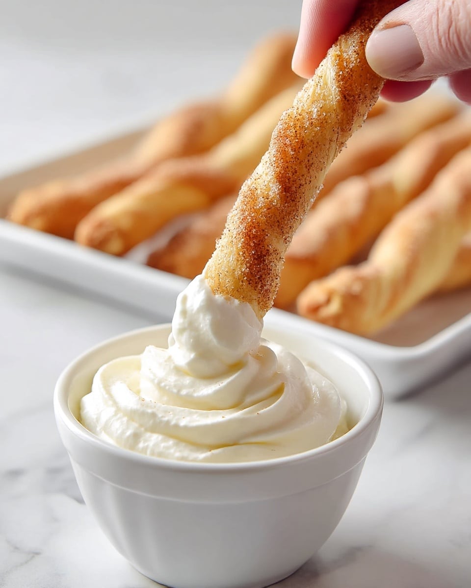 A close-up view shows a woman's hand dipping a twisted, golden-brown, cinnamon-sugar coated pastry stick into a swirl of fluffy white whipped cream in a small white bowl. In the blurred background, more of the same twisted pastry sticks lie in a row on a tray. The white bowl sits on a surface with a white marbled texture. The pastry stick has a crispy texture with visible sugar and cinnamon specks evenly spread on its surface. photo taken with an iphone --ar 4:5 --v 7