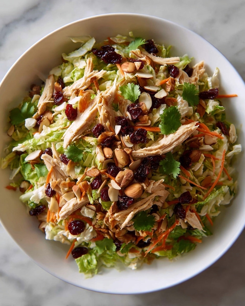 A white bowl filled with a layered salad starting with a base of light green shredded lettuce and finely sliced cabbage. On top, there are thin orange carrot strips and shredded pieces of cooked light brown chicken scattered evenly. Dark red dried cranberries and sliced almonds are sprinkled throughout, adding texture and color contrast. Small green cilantro leaves are spread across the salad, with some whole almonds and sesame seeds also visible. The bowl sits on a white marbled surface, and the overall texture looks fresh and crunchy. photo taken with an iphone --ar 4:5 --v 7