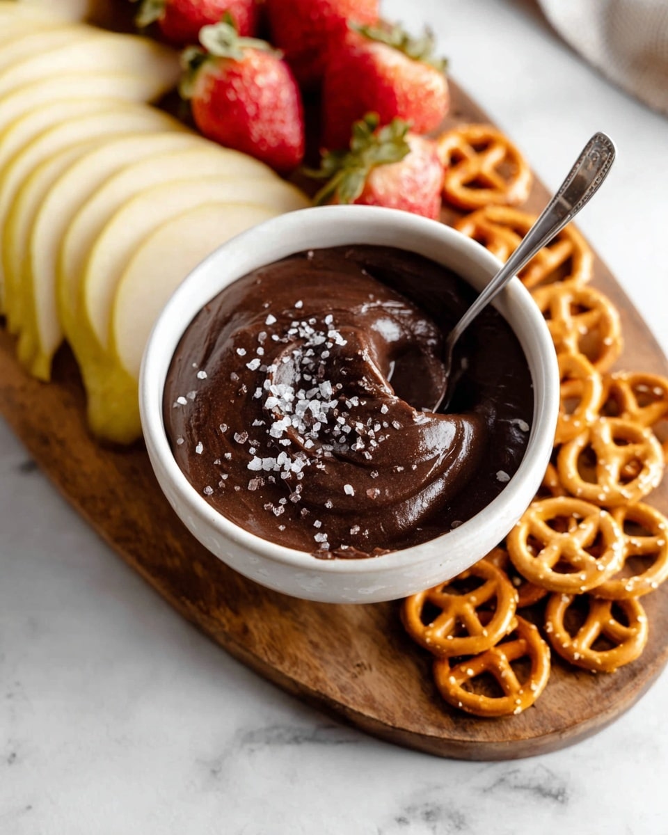 A white round bowl filled with smooth, dark chocolate dip topped with coarse salt crystals swirled on the surface, surrounded by four groups of dippables on a white marbled texture: thin light yellow apple slices on the top left, small golden pretzels on the top right, square light brown graham crackers at the bottom left, and bright red strawberries cut in halves on the bottom right, all arranged closely around the bowl. photo taken with an iphone --ar 4:5 --v 7