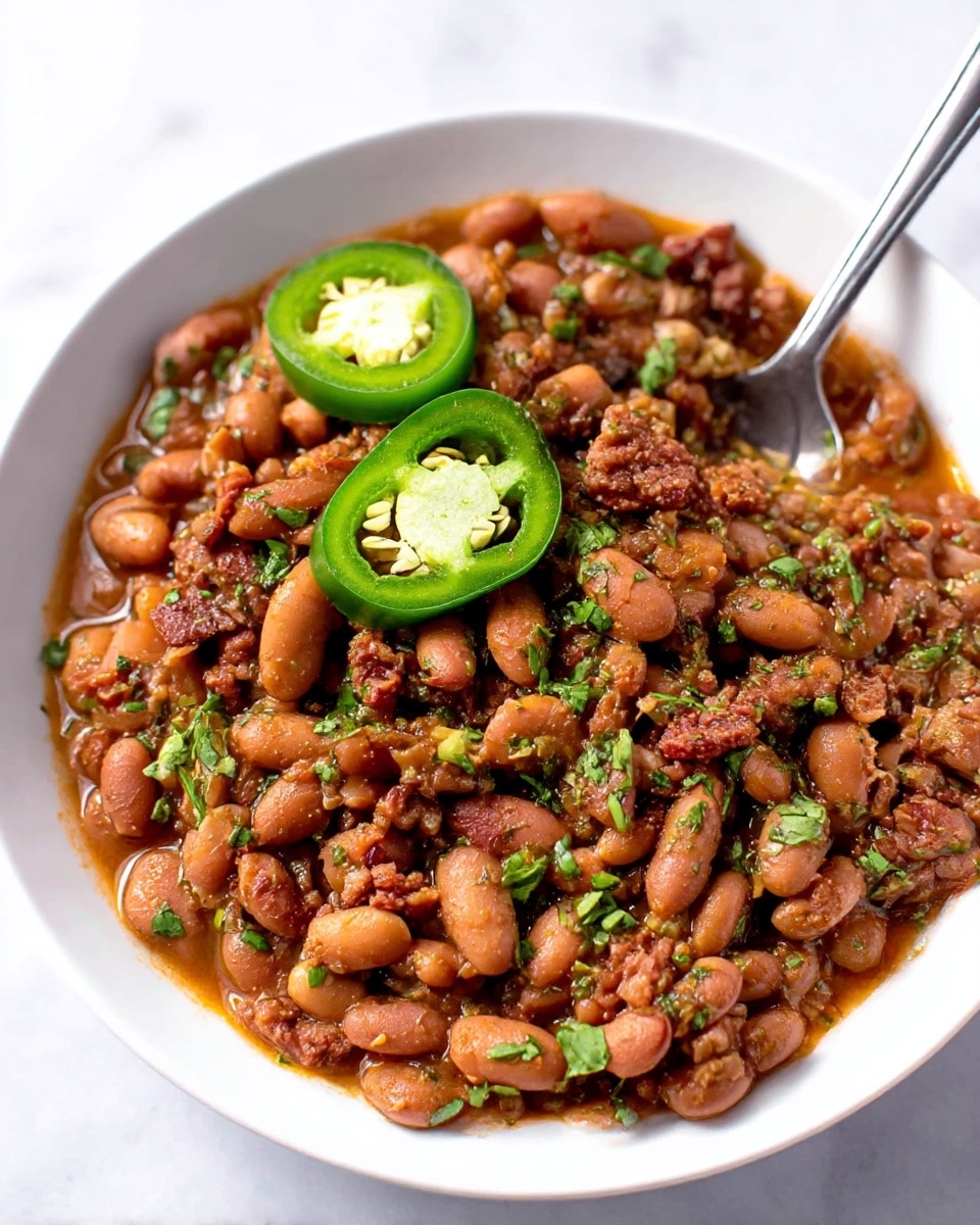 A white plate holds three layers of food arranged side by side: on the left are three yellowish-green slices of cooked zucchini with light grill marks and some green herbs sprinkled on them; in the middle is a mound of plain white rice with soft, fluffy texture; on the right is a generous portion of reddish-brown beans mixed with small pieces of cooked bacon and herbs, topped with two slices of bright green jalapeño pepper. A spoon rests at the bottom left of the plate, and the plate sits on a white marbled surface with part of a sliced avocado and a bowl of more beans blurred in the background. photo taken with an iphone --ar 4:5 --v 7