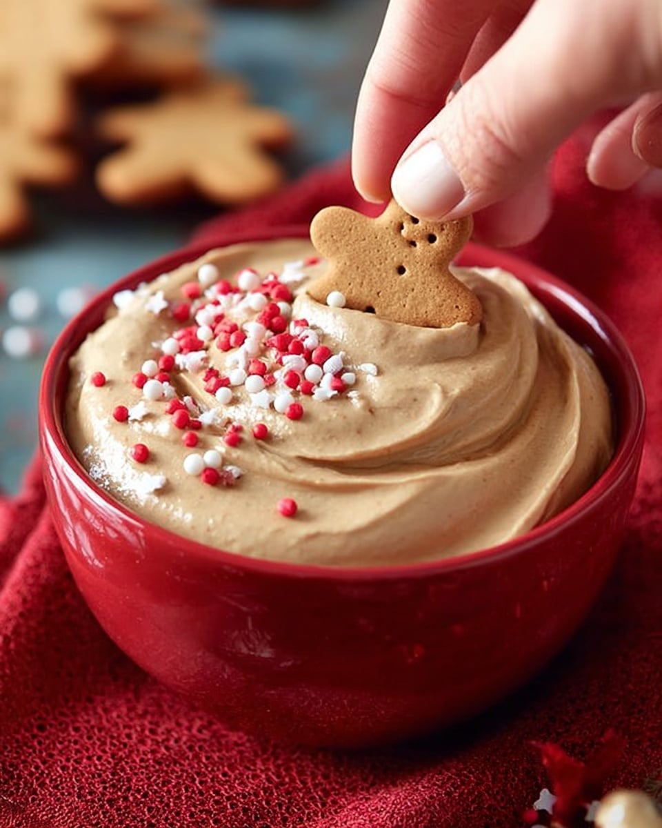 A red bowl filled with thick, light brown whipped cream or mousse shows smooth swirls on top, decorated with small red round sprinkles and gingerbread man-shaped white and brown sprinkles scattered across the surface. A woman's hand is dipping a small biscuit with a golden-brown color into the whipped cream. The bowl sits on a red cloth, which is placed on a textured surface. The background is softly blurred. The scene is bright and focused on the creamy dessert. Photo taken with an iphone --ar 4:5 --v 7