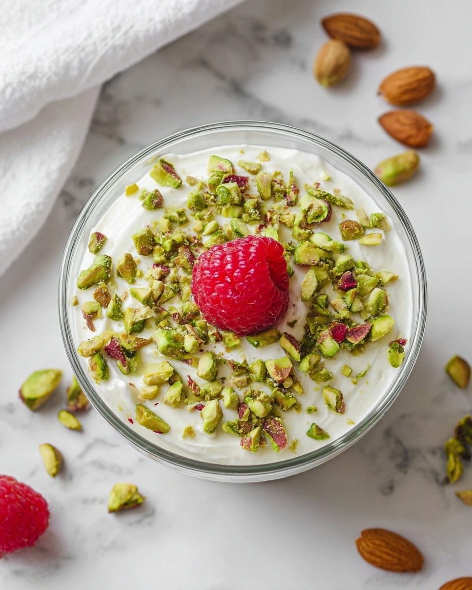 A clear round glass cup holds a two-layer dessert placed on a white marbled surface. The bottom layer is thick and creamy with a light green color speckled with tiny black chia seeds, showing a smooth and slightly chunky texture. The top layer is a smooth, white cream that covers the green base evenly. On top, there are many chopped pistachio nuts scattered around a single large, bright red raspberry placed in the center. Around the cup, there are a few whole pistachio nuts on the white marbled surface with a small white bowl partially visible in the background. photo taken with an iphone --ar 4:5 --v 7