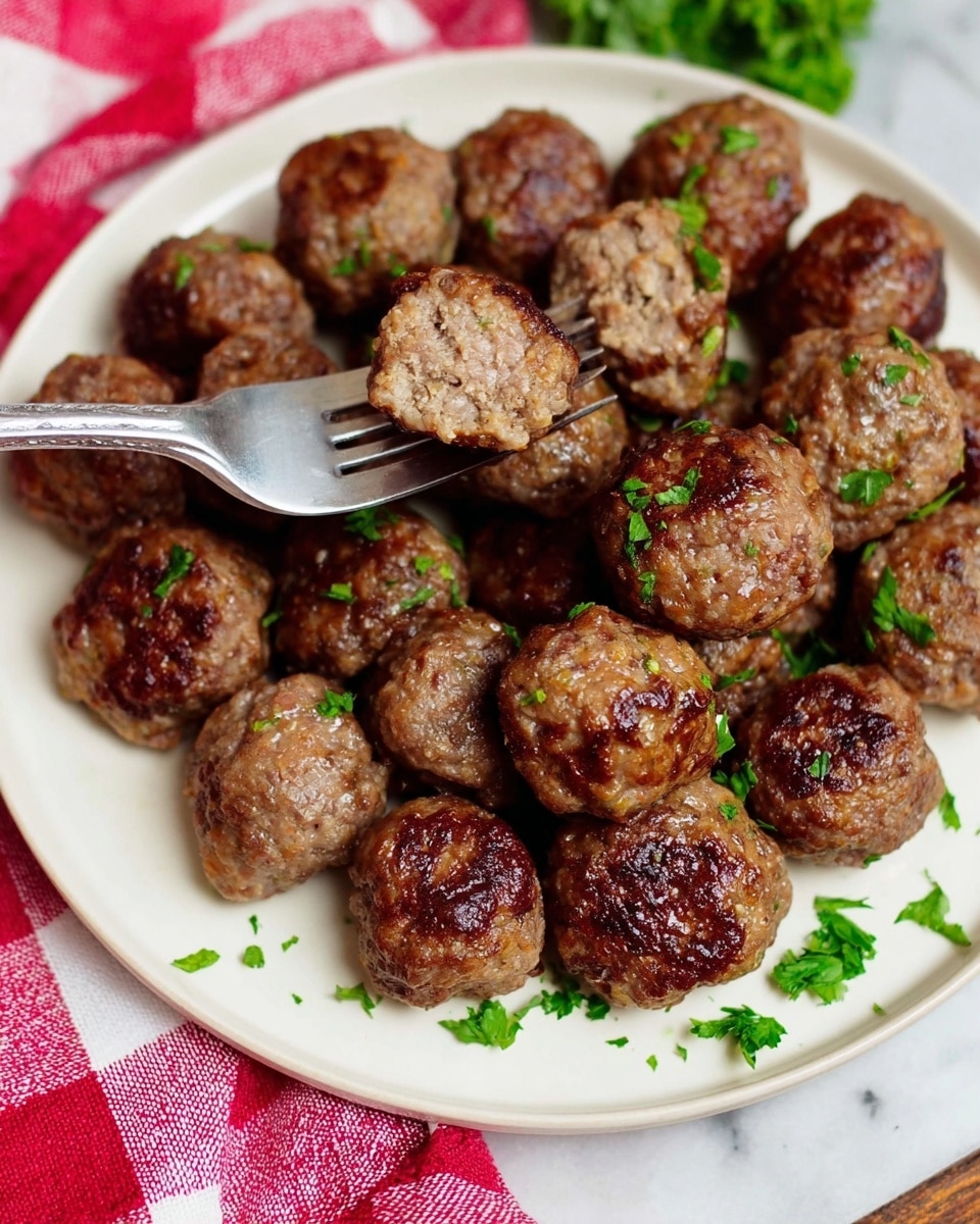 A white plate holds many round meatballs with a browned, slightly crispy outer layer and a tender, juicy inside visible in one meatball lifted by a fork from the left side. The meatballs are garnished with small green parsley pieces scattered on and around them. The plate rests on a white marbled surface with a red and white checkered cloth partially visible at the bottom left. Photo taken with an iphone --ar 4:5 --v 7
