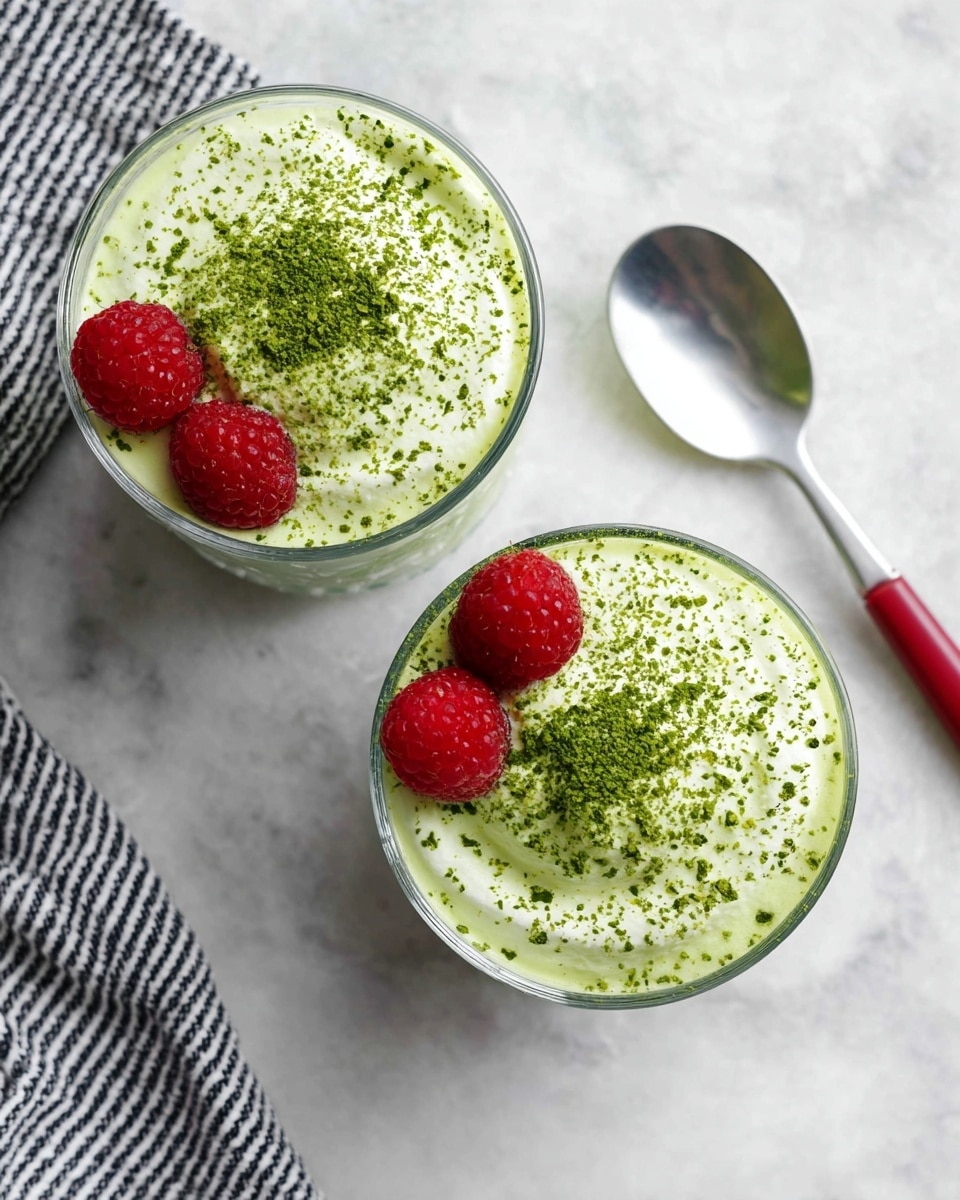 Two clear glass cups filled with a creamy light green drink, each topped with a smooth foam layer sprinkled with bright green matcha powder. On top of the foam in each cup, there are two fresh red raspberries positioned close together. The cups sit on a white marbled surface, with a silver spoon with a red handle to the right, and a black and white striped cloth partially visible on the upper left side. The overall look is fresh and inviting, with a clean and bright setting. photo taken with an iphone --ar 4:5 --v 7