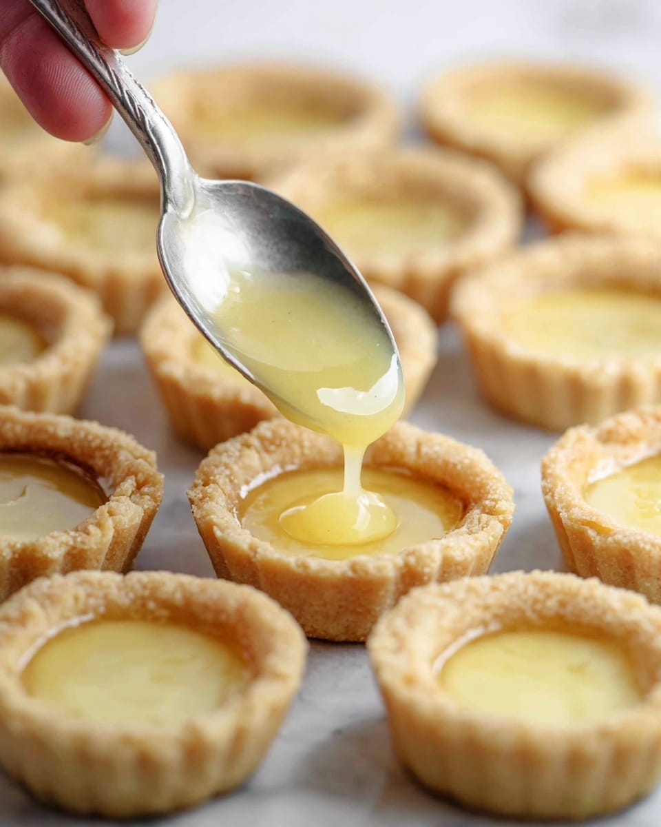 The image shows many small, round tart shells arranged close together on a white marbled surface. Each shell is light golden brown with a slightly crumbly texture, forming the base layer. A woman's hand holds a silver spoon filled with a glossy pale yellow filling that is being poured into one of the tart shells, creating a smooth, shiny second layer inside the shell. The focus is on the spoon and the tart shell being filled, with the other empty and partially filled shells blurred slightly in the background. photo taken with an iphone --ar 4:5 --v 7
