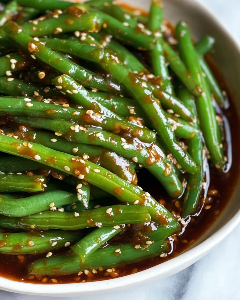 A close-up view of a dish featuring bright green whole green beans covered in a thick, glossy dark reddish-brown sauce with visible sesame seeds scattered all over. The green beans are fresh and shiny with a slight gloss from the sauce, which pools slightly at the bottom, adding a rich texture. The dish is served in a white bowl, and the background shows a white marbled texture. Photo taken with an iphone --ar 4:5 --v 7