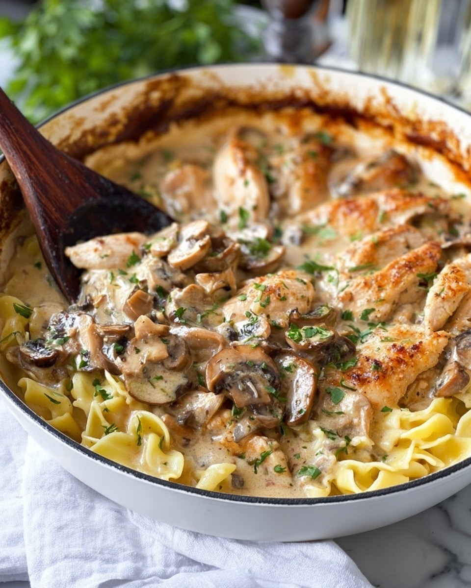 A white pan filled with a creamy chicken and mushroom dish showing two main layers: the bottom layer with light yellow pasta and large pieces of cooked chicken in a golden-brown color, and the top layer with sliced mushrooms and finely chopped green herbs mixed in a thick, pale cream sauce. The edges of the pan show browned, slightly crispy spots from cooking. A dark wooden spatula is partially inside the pan, resting on the edge, and there is a white cloth under the pan on a white marbled surface with some green leaves blurred in the background. photo taken with an iphone --ar 4:5 --v 7