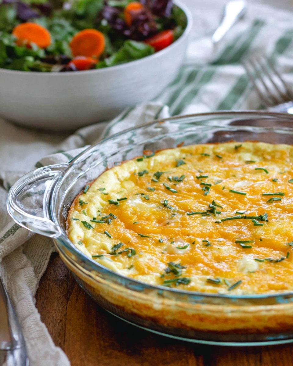 A round baked dish in a clear glass pie pan with two side handles rests on a wooden table with a white marbled texture underneath. The dish has a golden-yellow top layer of melted cheese with lighter creamy spots, sprinkled evenly with small, bright green herb pieces. In the background, there is a blurred bowl of mixed salad with visible green leaves, orange carrot slices, and red tomato chunks. The setting includes a white cloth with a soft green striped detail and a shiny metal fork near the pie pan. photo taken with an iphone --ar 4:5 --v 7