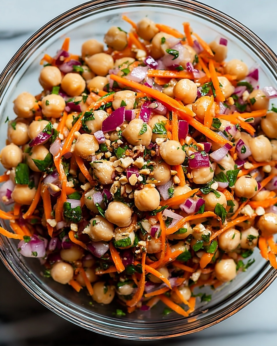 A close-up view of a clear glass bowl filled with a vibrant chickpea salad. The salad has three main layers: round, light beige chickpeas are the biggest part, mixed with bright orange thin carrot strips, and chopped purple-red onion pieces. Fresh green herbs are spread evenly throughout the bowl, adding pops of green color. Small bits of crushed nuts and a light dressing give the salad a shiny look. The bowl is set on a smooth white marbled texture. photo taken with an iphone --ar 4:5 --v 7