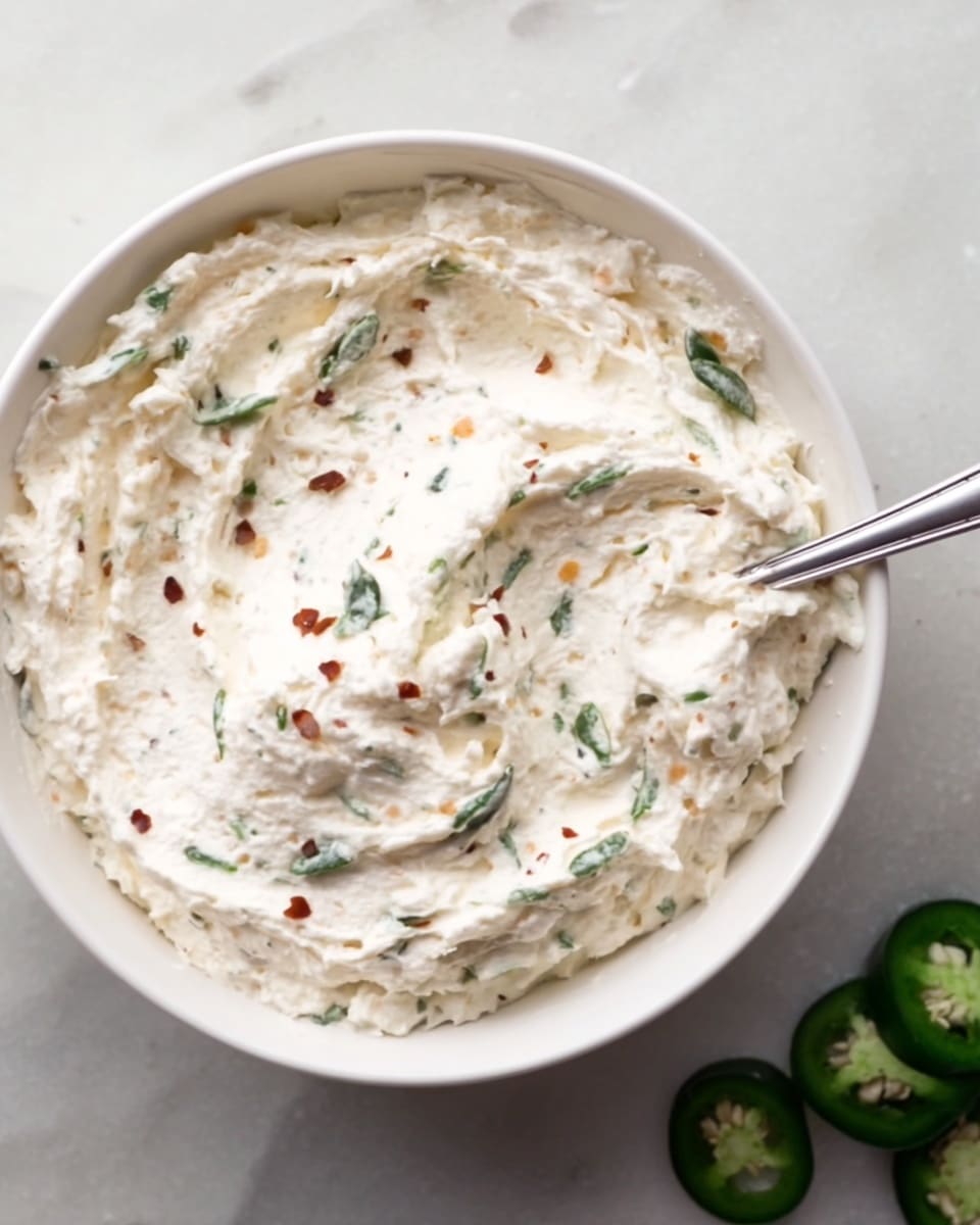 A white bowl filled with a creamy, thick white spread that has a slightly bumpy texture. The spread is mixed with small green leaves and tiny red flakes scattered throughout. A silver spoon is partially inside the bowl on the right side. The bowl is placed on a white marbled surface with two green jalapeño slices visible at the bottom right corner. Photo taken with an iphone --ar 4:5 --v 7