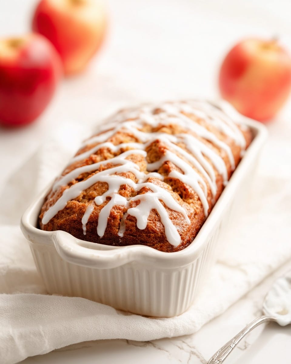 A golden-brown baked loaf with a textured top glistening slightly, drizzled with uneven, smooth white icing in a crisscross pattern, sits inside a white ceramic rectangular baking dish with rounded handles. The dish is resting on a soft white cloth over a white marbled surface. In the blurred background, there are a few red apples, adding a pop of color to the scene. A spoon with some white icing on it is partially visible near the front right corner. The overall look is clean, warm, and inviting. photo taken with an iphone --ar 4:5 --v 7
