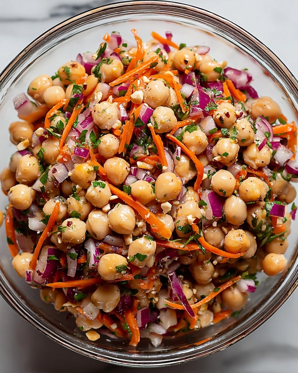 A clear glass bowl filled with a colorful chickpea salad consisting of creamy light beige chickpeas, thin bright orange carrot strips, small pieces of purple-red onions, and scattered green herbs. The chickpeas and vegetables are mixed together with small bits of crushed nuts and some shiny dressing that gives a lightly glossy texture. The bowl is placed on a white marbled surface, enhancing the vibrant colors of the salad. photo taken with an iphone --ar 4:5 --v 7