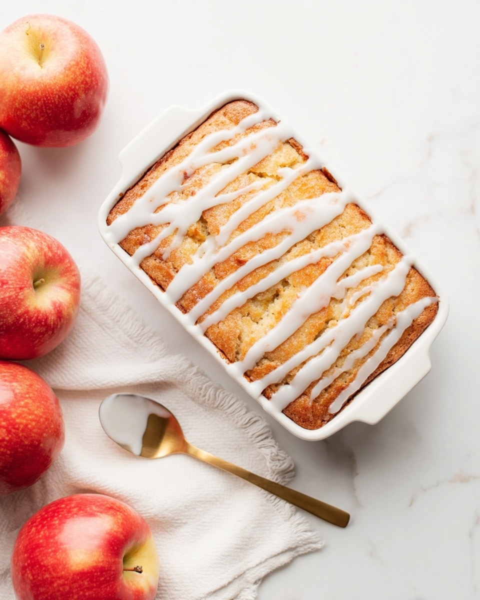 A golden-brown baked square cake in a white rectangular dish, topped with a white icing glaze drizzled in irregular lines across the surface. The cake has a slightly rough texture, showing it is soft on the inside. Next to the dish, there are three red apples with shiny skin, adding vibrant color to the scene. A gold spoon with some white icing on it rests on a white cloth napkin under the dish. The whole setting is on a white marbled surface, creating a clean and bright background. photo taken with an iphone --ar 4:5 --v 7