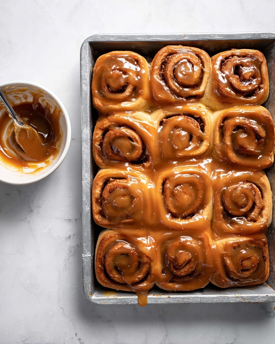 A metal tray filled with nine soft, thick cinnamon rolls arranged in three rows of three, each roll showing visible spiral layers of golden brown dough with darker cinnamon filling. The rolls are generously covered in a shiny, thick, caramel-colored glaze that drips slightly onto the tray. To the left of the tray is a small white bowl with leftover glaze and a spoon inside, sitting on a white marbled surface. photo taken with an iphone --ar 4:5 --v 7